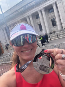 A woman in running gear smiles and holds up a Half Marathon medal